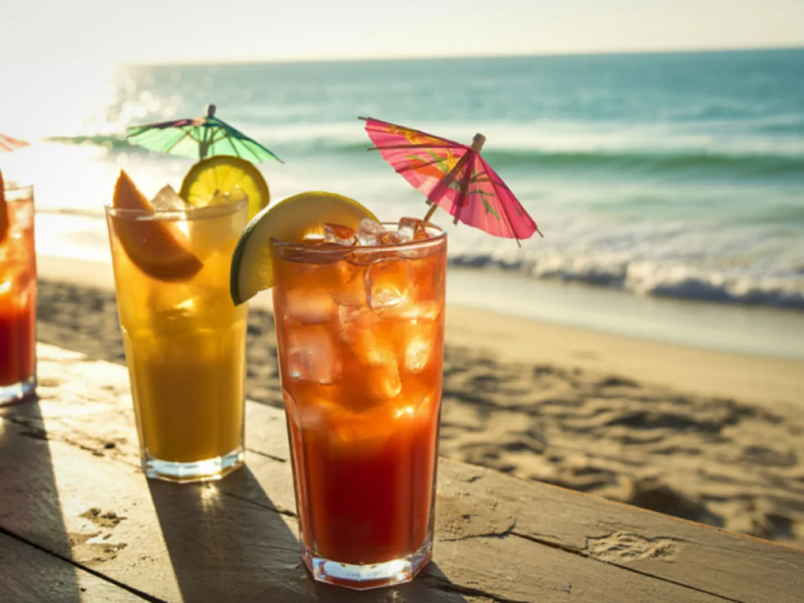 Three beach themed cocktails sitting on the sand at the beach.